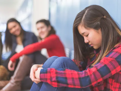 Three girls in the background talking amongst themselves and laughing at girl sitting by herself against the locker.